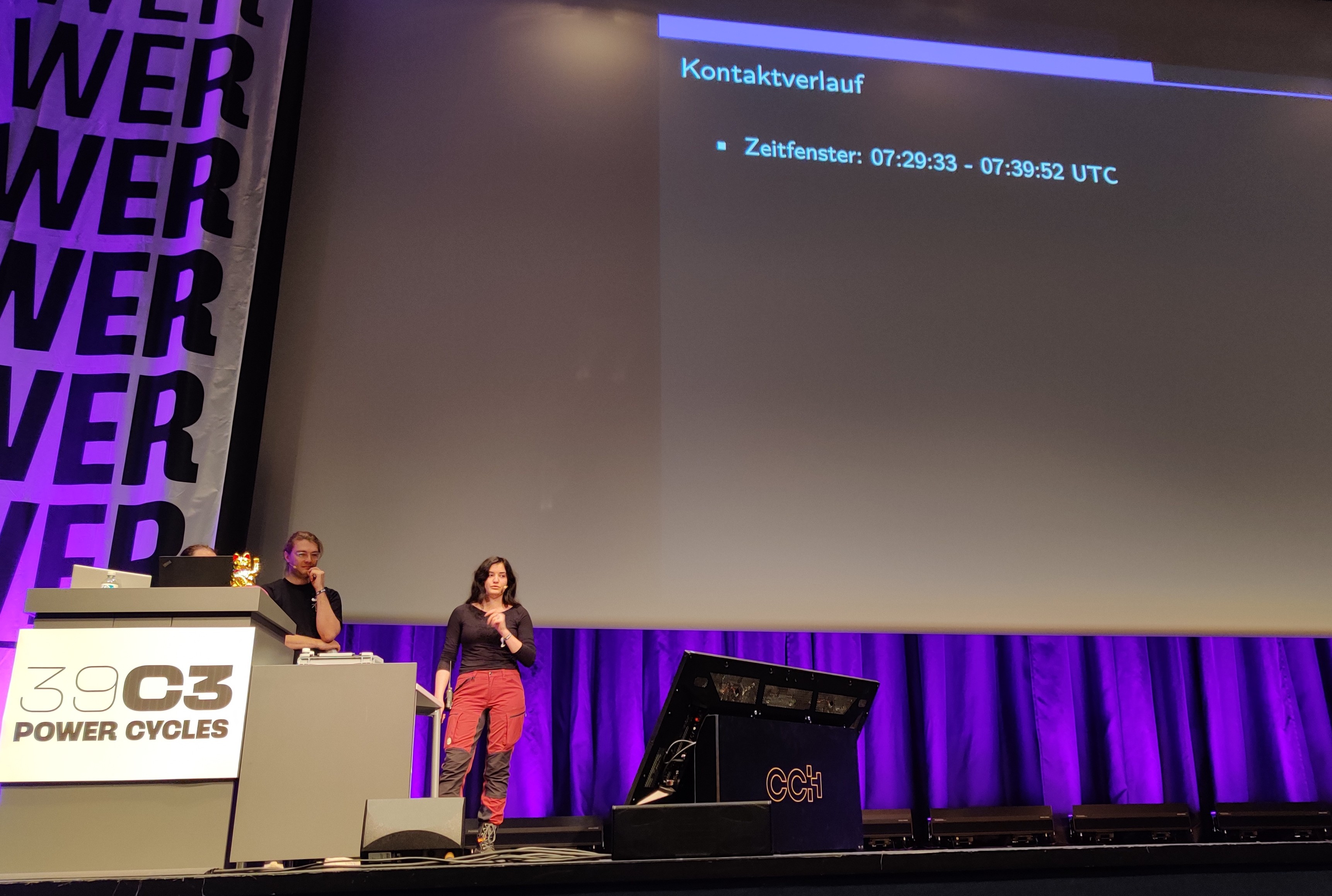 Dunkle Bühne mit Leinwand und Präsi-Folien im Hintergrund. Vorne links stehen eine Frau und zwei Männer und halten einen Vortrag